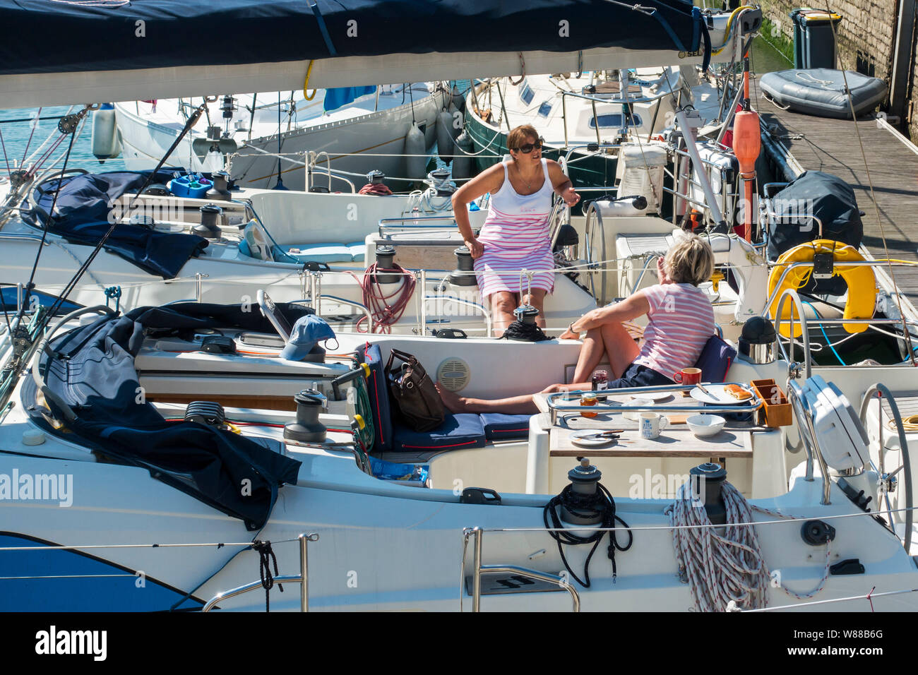 Wealthy women chatting after breakfast on board of their neighbouring yachts / sailing boats docked / moored in marina / yacht basin / port in summer Stock Photo
