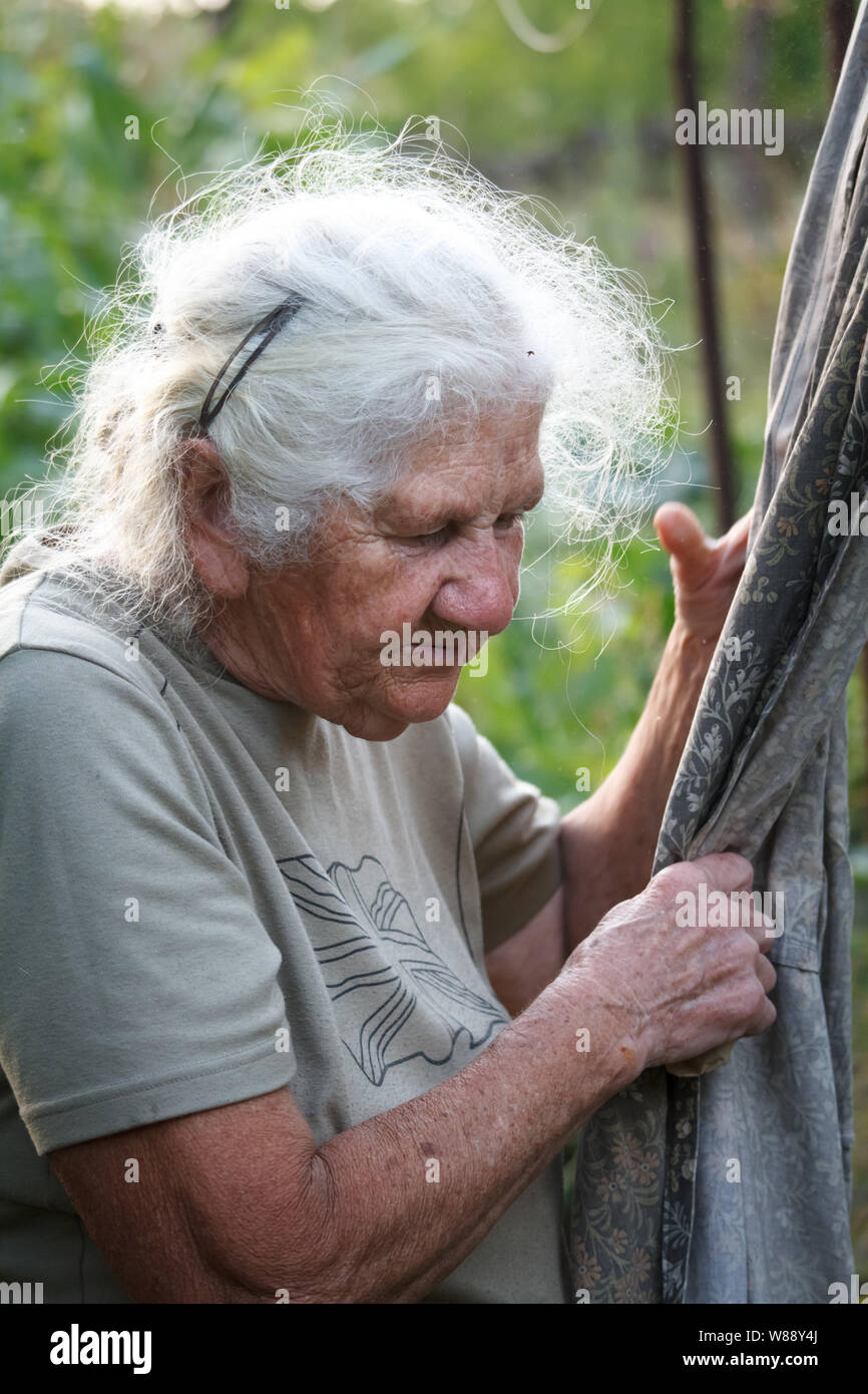 Close-up portrait of an old woman with gray hair hangs clothes to dry on a blurred background of a green meadow, selective focus Stock Photo