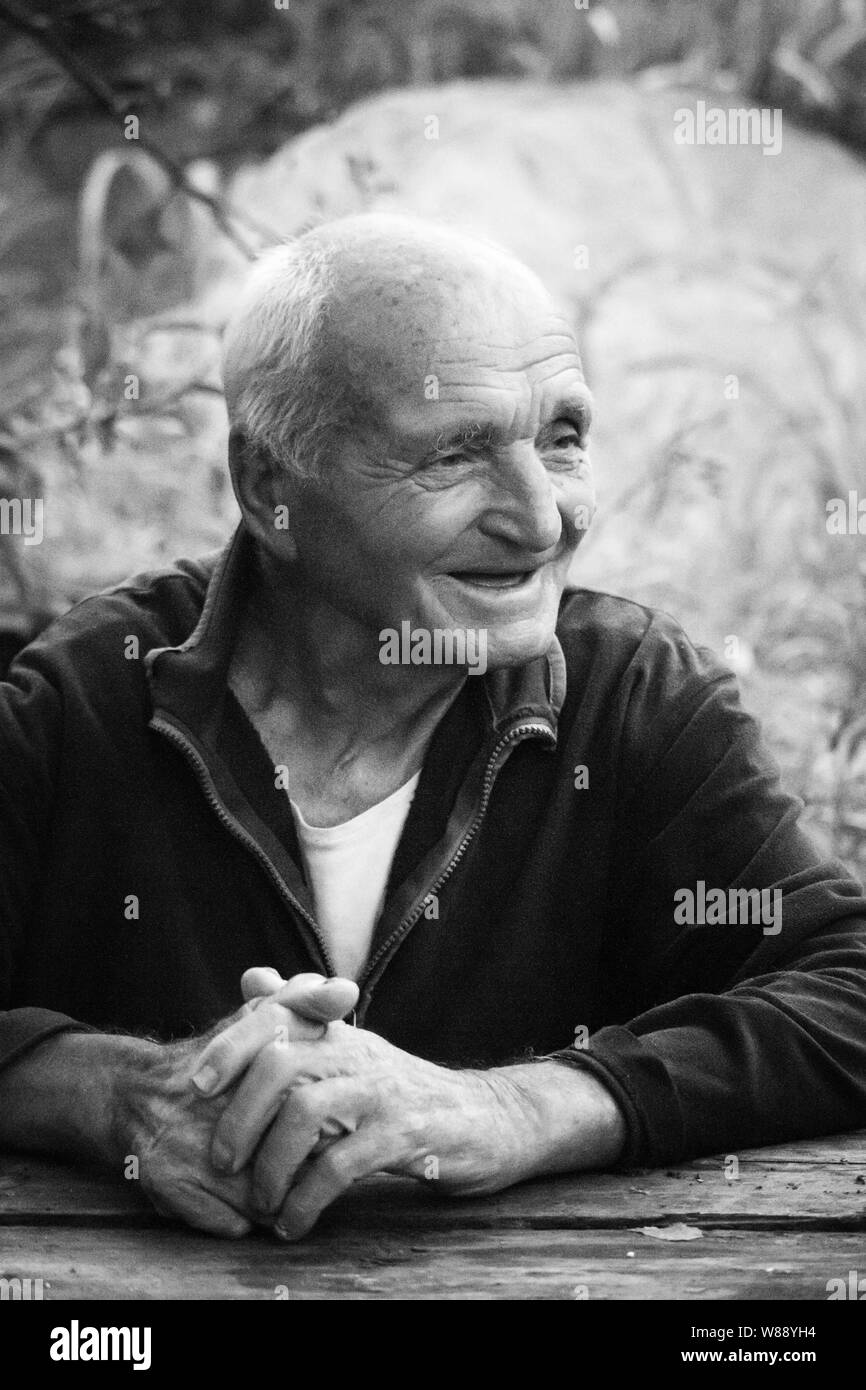Closeup black white portrait of a very old man with arms crossed, sitting at a table on a blurred background of green trees, selective focus Stock Photo