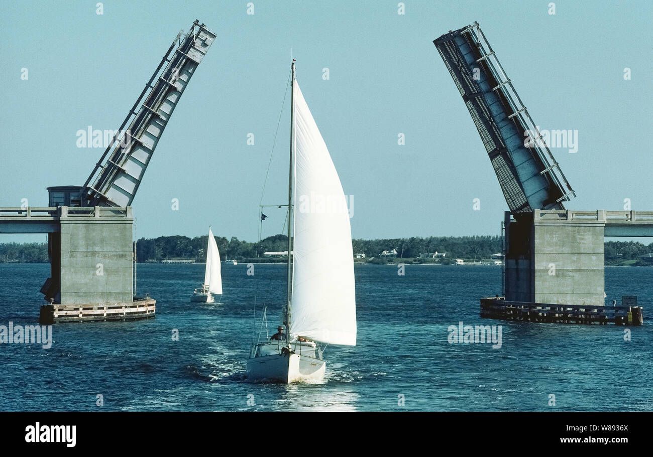 A double-leaf drawbridge opens to allow two sailboats to continue their passage on the Indian River near St. Lucie on the east coast of Florida, USA. Technically known as a bascule bridge (from the French word for seesaw), it has two identical steel lengths that span the waterway and open into the air with the aid of counterweights in the concrete support piers on either side. The design of this movable bridge is similar to those that spanned the moats of medieval castles, except they were single-leaf types that opened into the air only from one side. Stock Photo