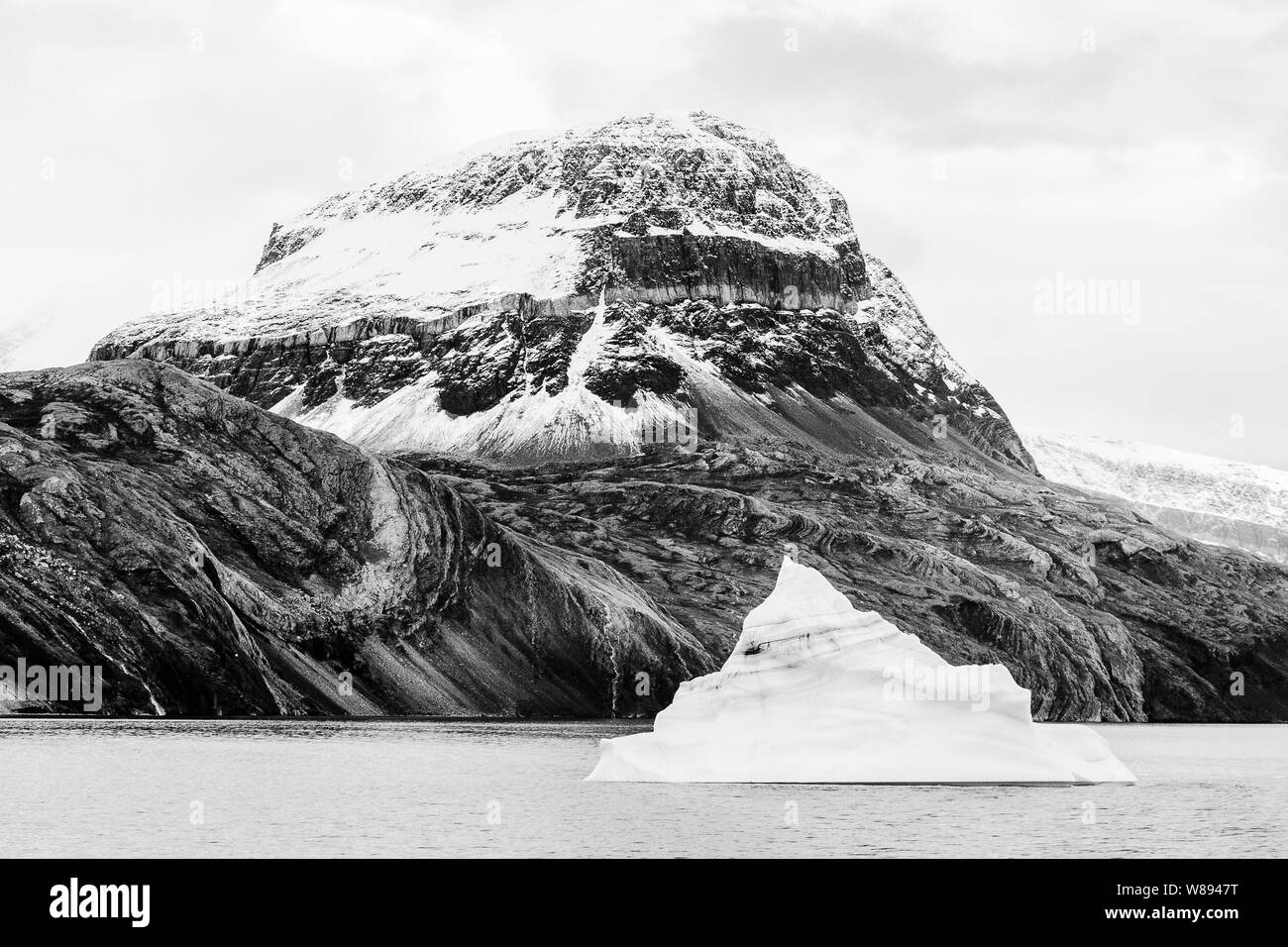 A cone shaped iceberg is dwarf by massive mountain slopes in Kejser Franz Joseph Fjord, Greenland Stock Photo