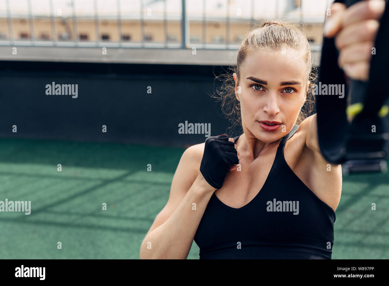 Young woman doing exercises on a rooftop. Fitness female working out outdoors using suspension strap for lifting. Stock Photo