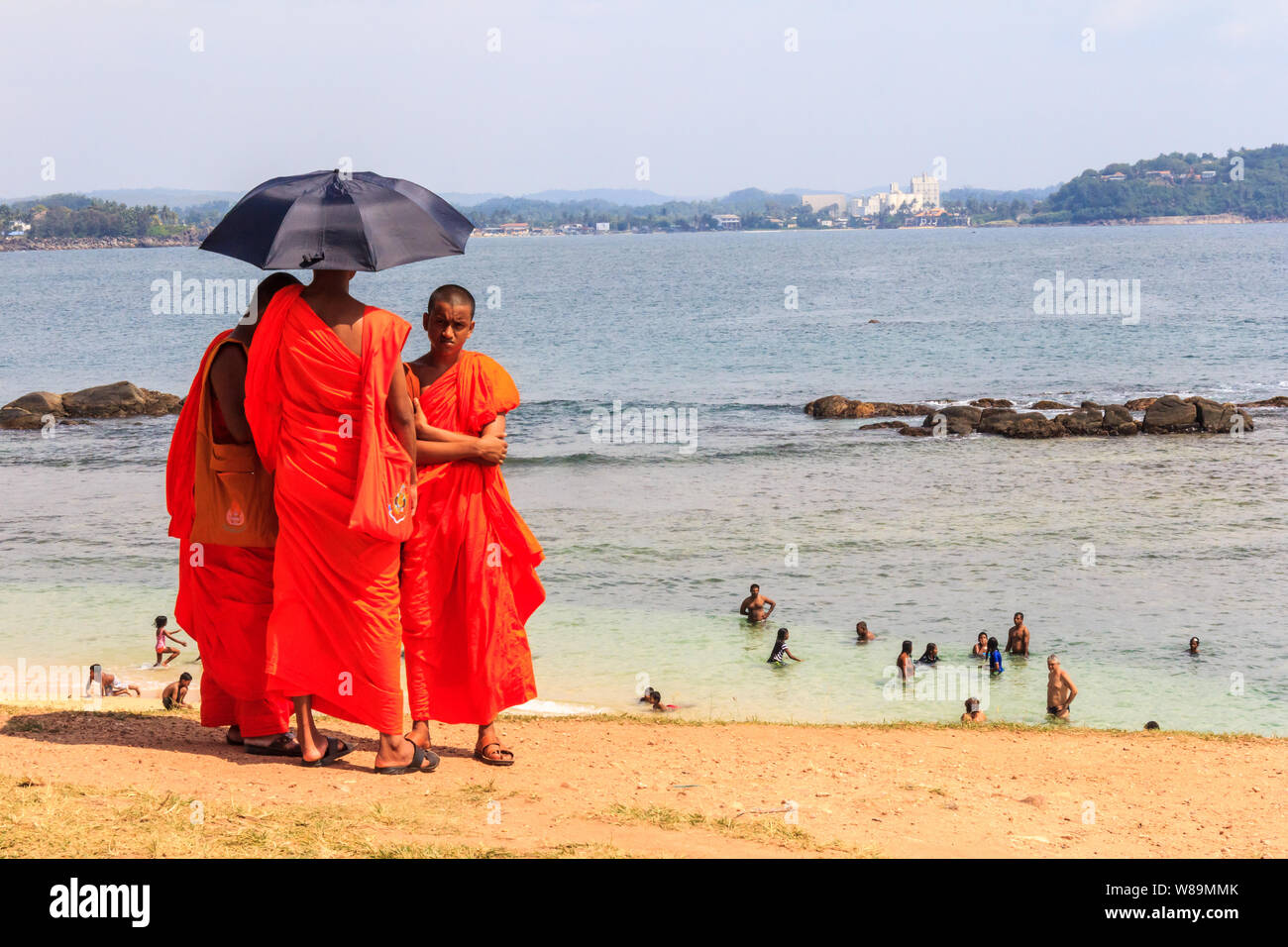 Galle, Sri Lanka - 30th December 2016: Monks shelter under an umbrella while people swim in the sea, The heat can be oppressive. Stock Photo
