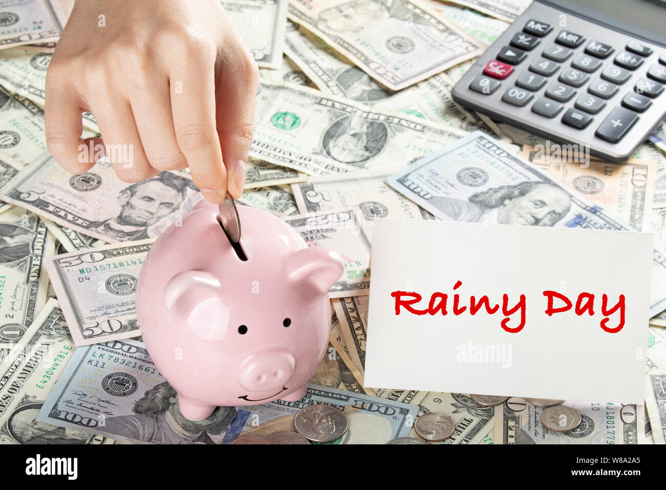 United States bank notes and coins with a calcultor, piggy bank, and hand placing coin into piggy bank and a card that says Rainy Day Stock Photo