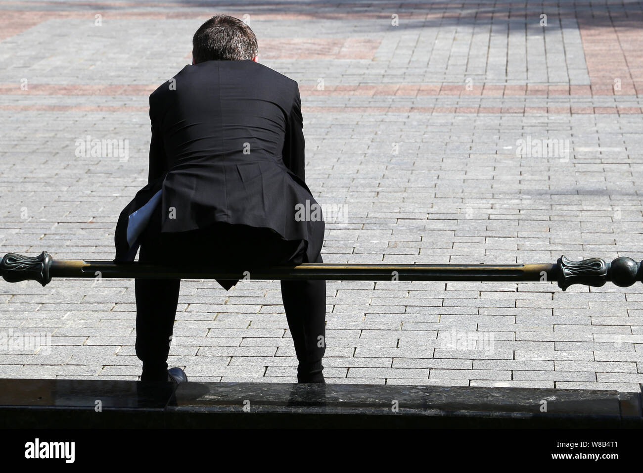 Man in a black business suit sitting on a railing on a city street, rear view. Concept of businessman, dismissed office worker, official, government Stock Photo