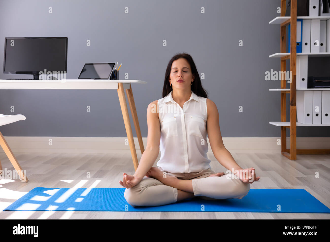 Young Businesswoman Sitting On Blue Exercise Mat Meditating In Office Stock Photo