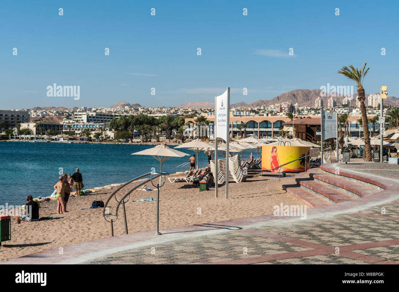 Eilat, Israel- November 7, 2017:   City promenade with palm trees and beach equipped with sunbeds and umbrellas. In the background city buildings and Stock Photo