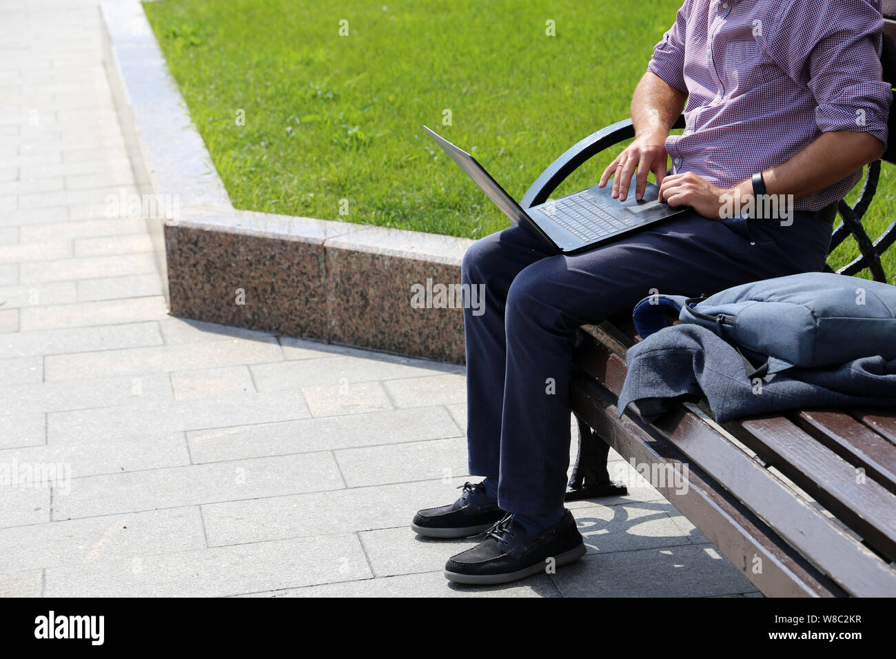 Man sitting with a laptop on a bench in a summer city on green lawn background. Male hands on a keyboard, concept of businessman, working outdoor Stock Photo