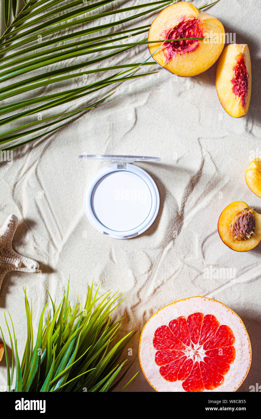 Dry cosmetic decorative product in a round refile lies on the sand, surrounded by bright fruits, corals, and palm leaves. Powder, highlighter, bronzer. Stock Photo