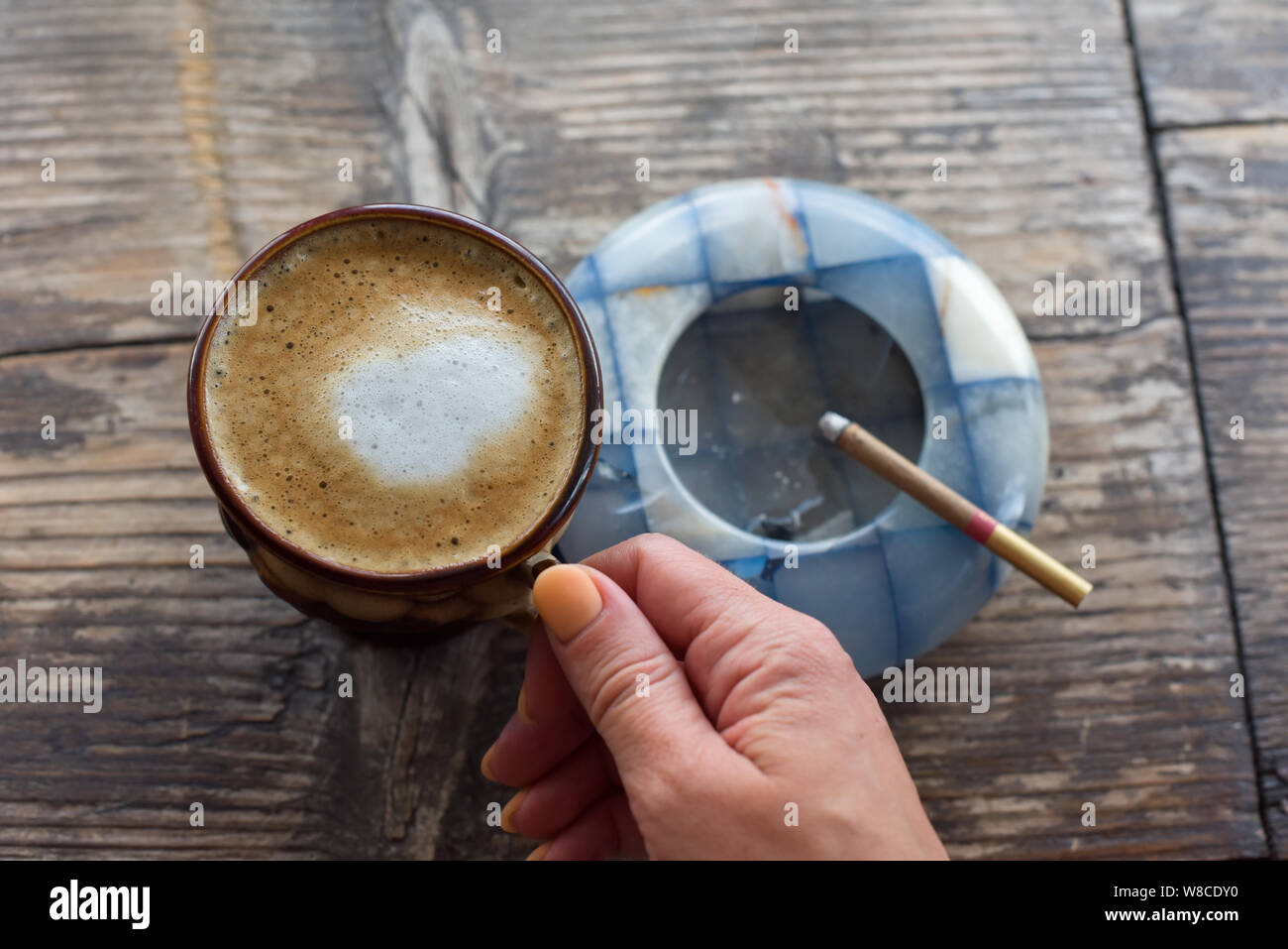 cappuccino cigarettes Coffee cigarette on the table. Cigarette in a woman's hand with a beautiful  manicure. Blue ashtray. The smoke of cigarettes. Brown cup with coffee on  Stock Photo