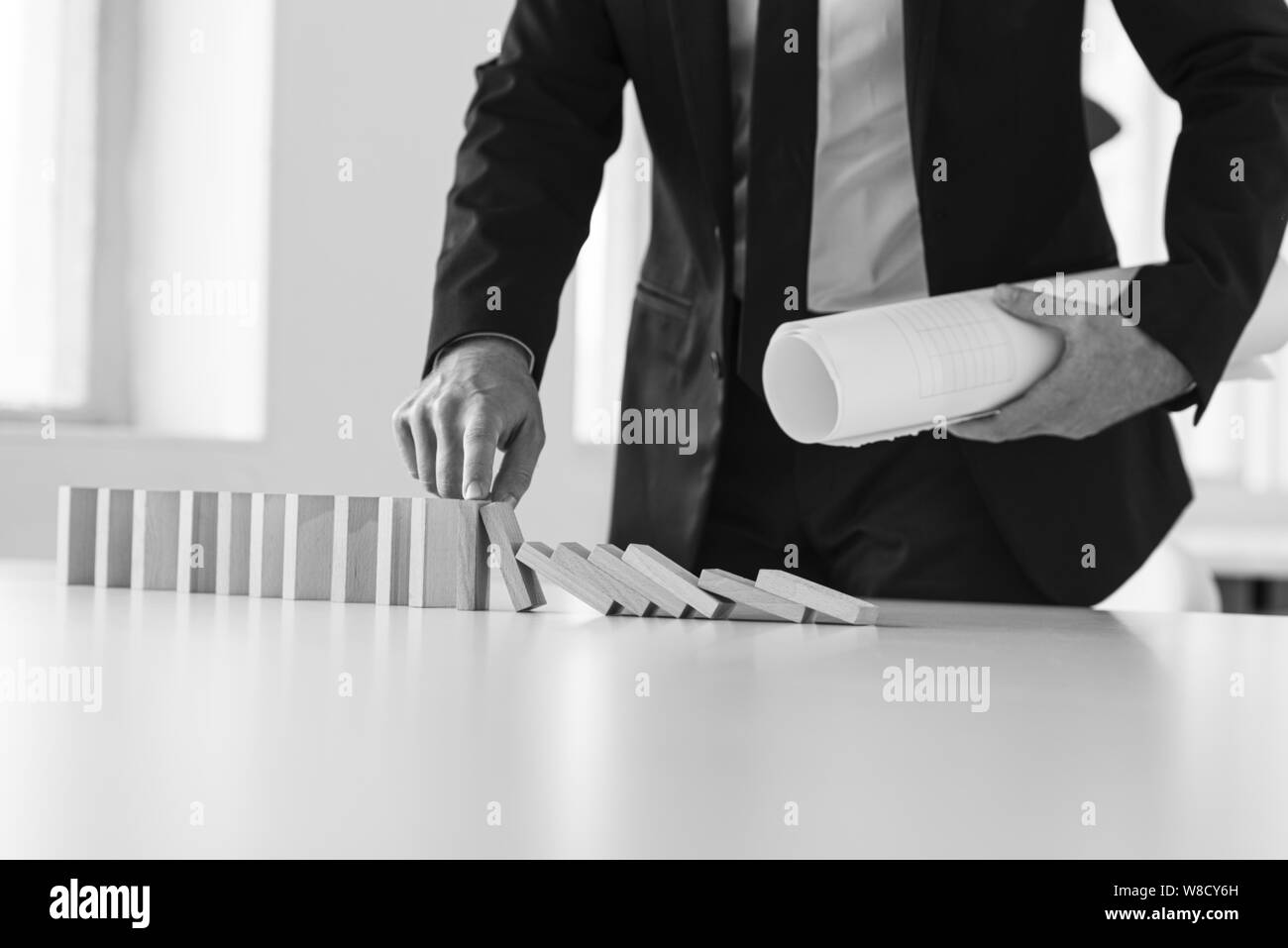Greyscale image of businessman holding rolled documents or project plans stopping falling dominos with his finger. Stock Photo