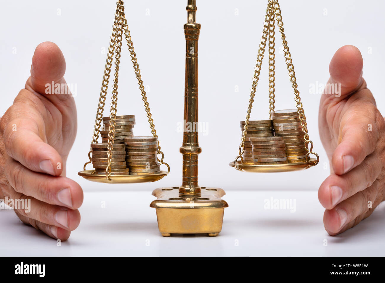 Close-up Of A Businessperson's Hand Covering Stacked Coins On Golden Weighing Scale Stock Photo