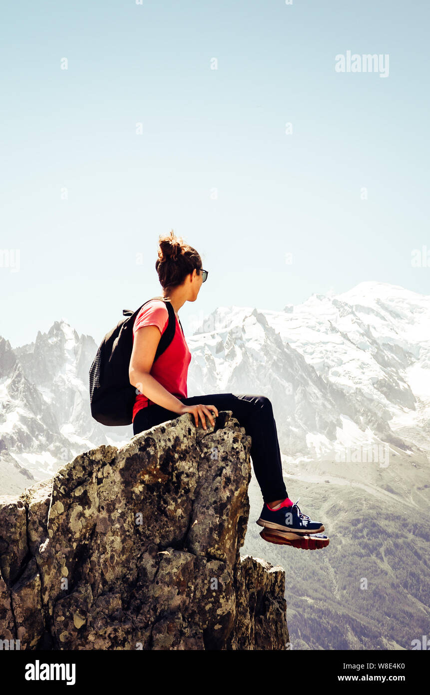 Vertical picture of young female traveller sitting on the edge of rocks. High mountains in background. Woman backpacking. Hiking adventure. Active lif Stock Photo