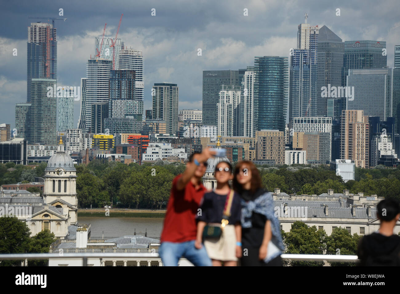 People take selfies in front of the Old Royal Naval College and Canary Wharf, from the Royal Observatory in Greenwich, London. Warnings for rain and wind came into force across nearly all of the UK today. Stock Photo