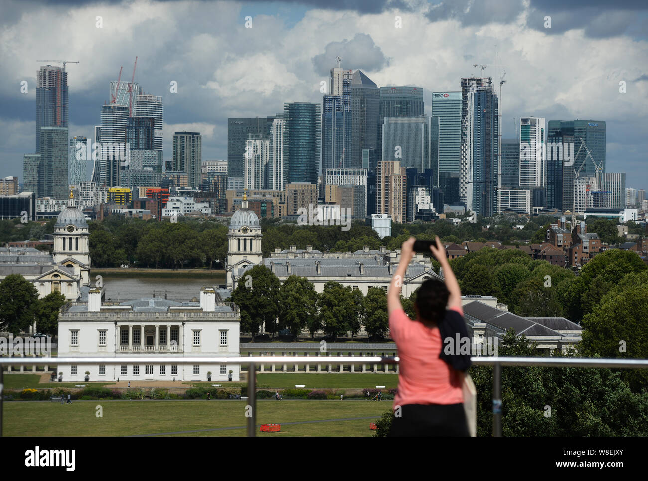 A woman takes a photograph of the Old Royal Naval College and Canary Wharf, from the Royal Observatory in Greenwich, London. Warnings for rain and wind came into force across nearly all of the UK today. Stock Photo