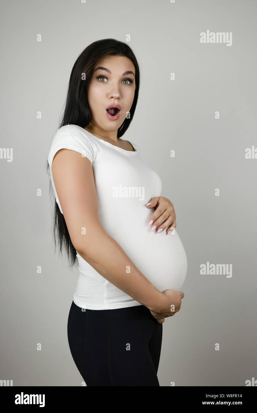 Beautiful Pregnant Brunette Woman Holding Her Pregnant Belly With Surprise On Isolated White