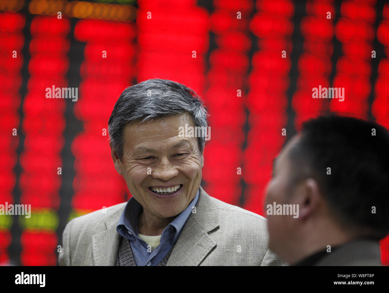 A Chinese investor smiles and talks to another in front of a display showing prices of shares (red for price rising and green for price falling) at a Stock Photo