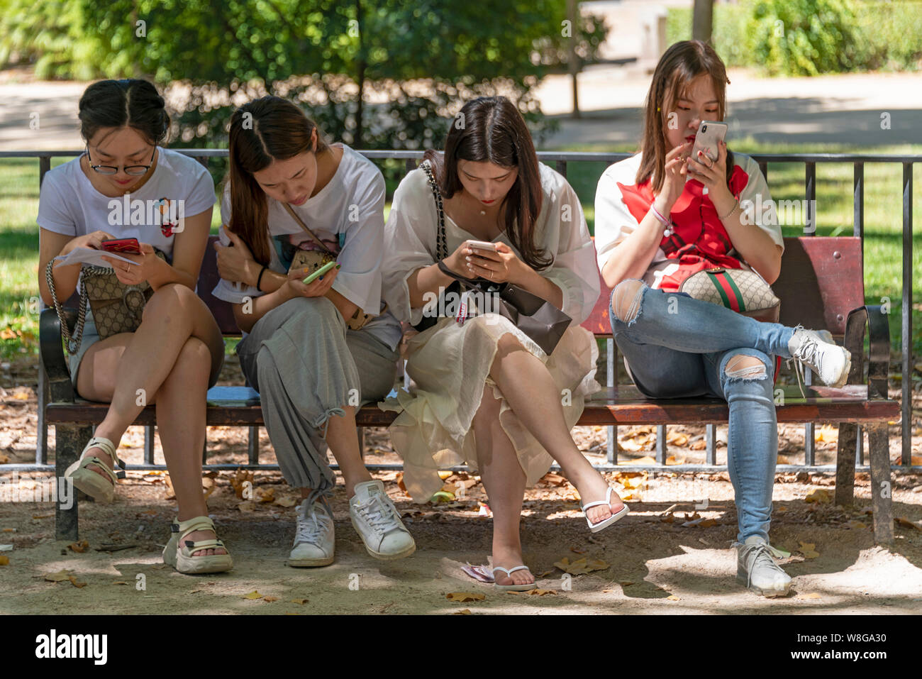 Horizontal portrait of four Asian friends sitting together on a park bench using their phones. Stock Photo