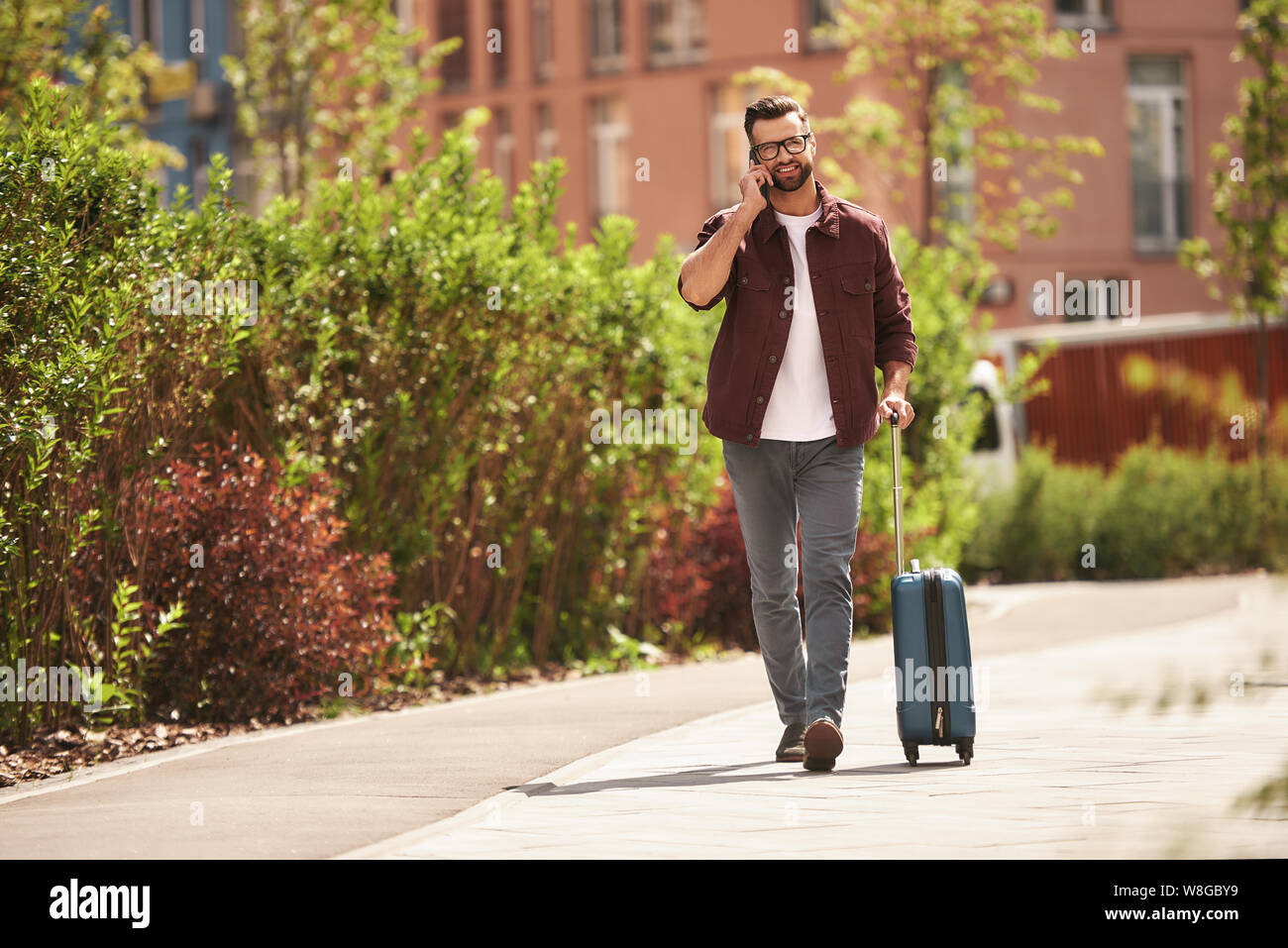 Happy to hear you Cheerful bearded man in casual wear and eyeglasses pulling his luggage and talking by phone while walking through the city street. Travel concept. Lifestyle Stock Photo