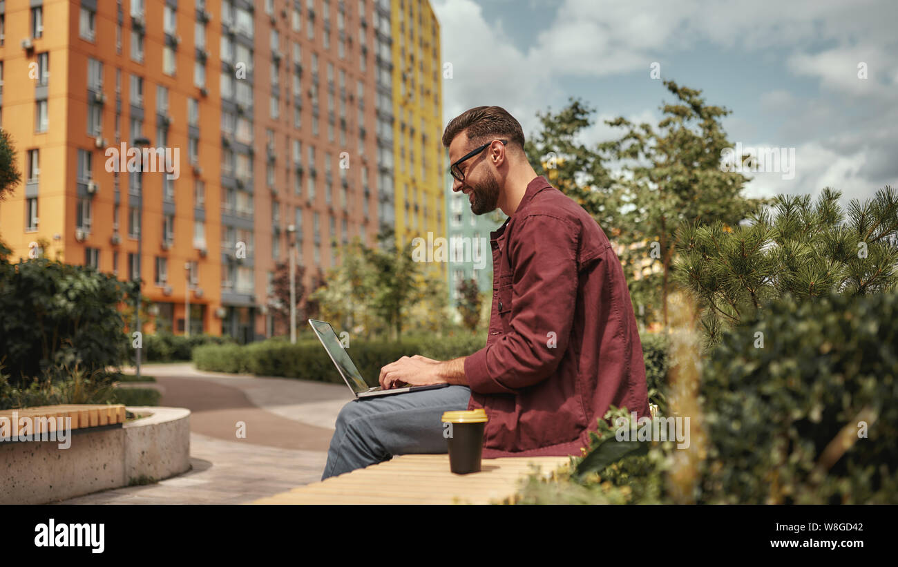 Happy and young. Side view of handsome man with stubble in casual clothes and eyeglasses working on laptop while sitting on the bench outdoors. Blogging. City life. Work Stock Photo