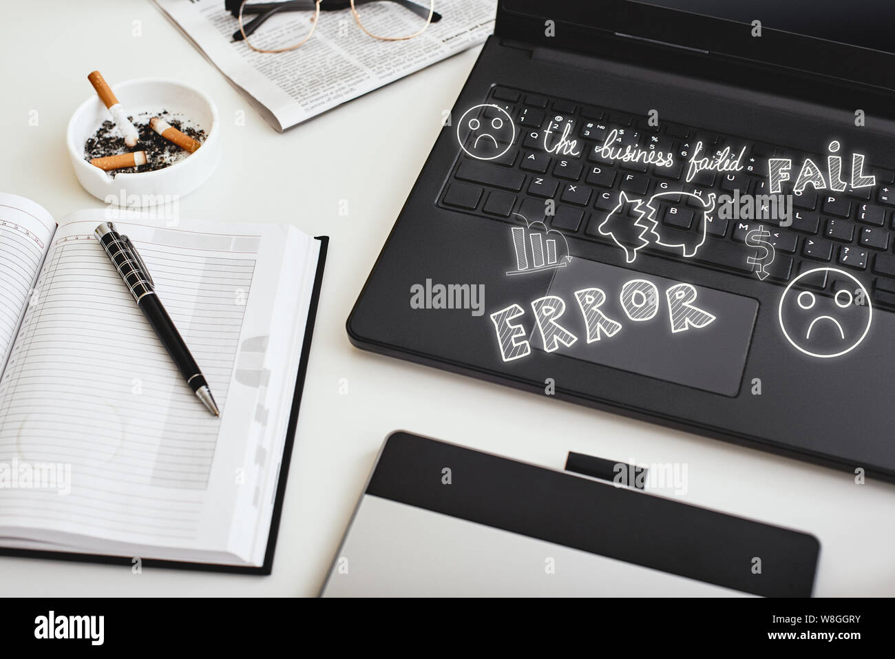 A laptop, pen, ashtray with cigarette ends, glasses and notebook lying
