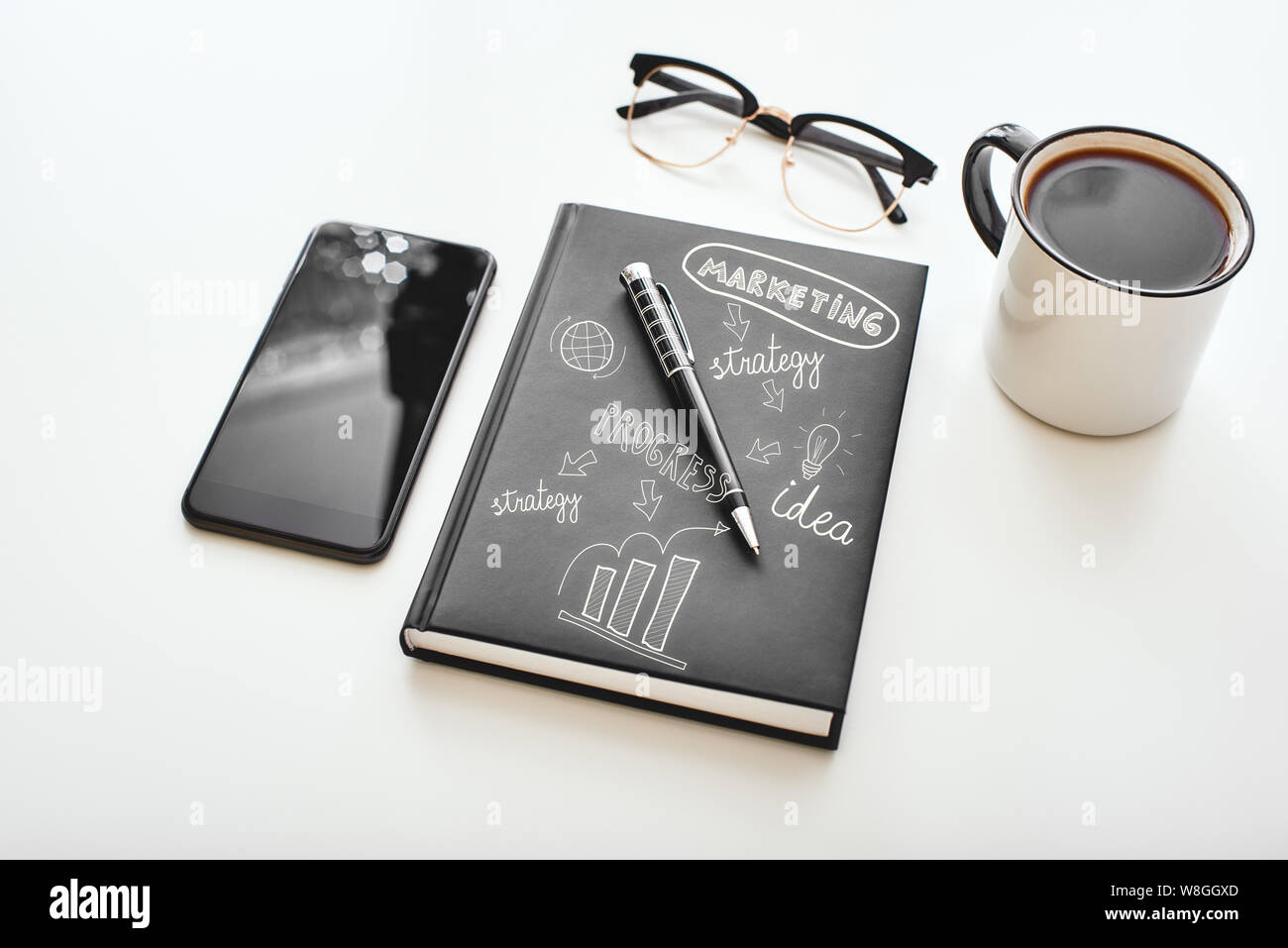 Isolated cup of coffee, smartphone, pen, notebook and glasses lying on a white desk. Hand drawn doodle illustration on the notebook. Horizontal shot Stock Photo