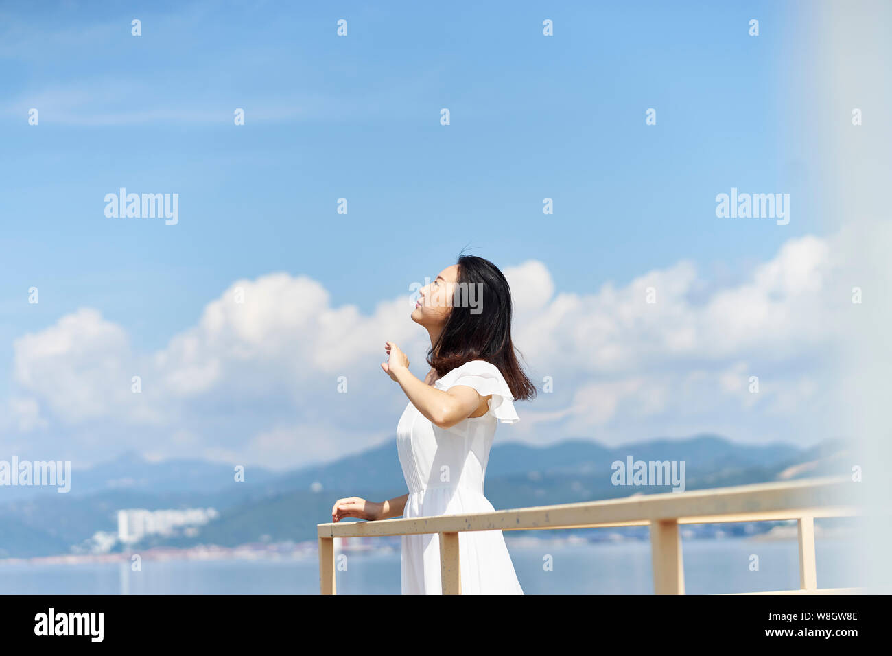 side view of a young asian woman standing by sea looking up at blue sky Stock Photo