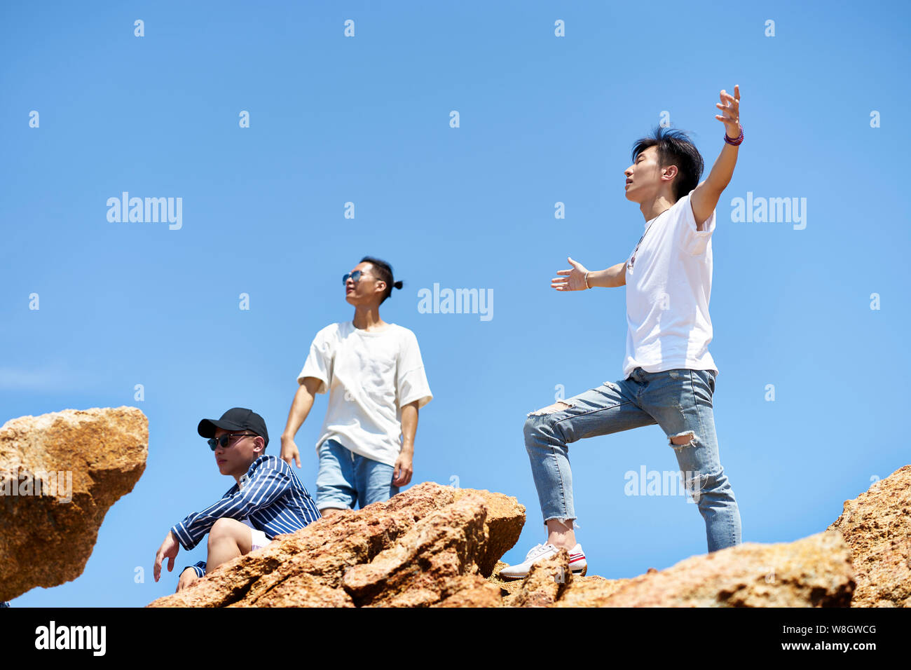 group of young asian adult men standing on top of rocks against blue sky enjoying sunshine and fresh air Stock Photo