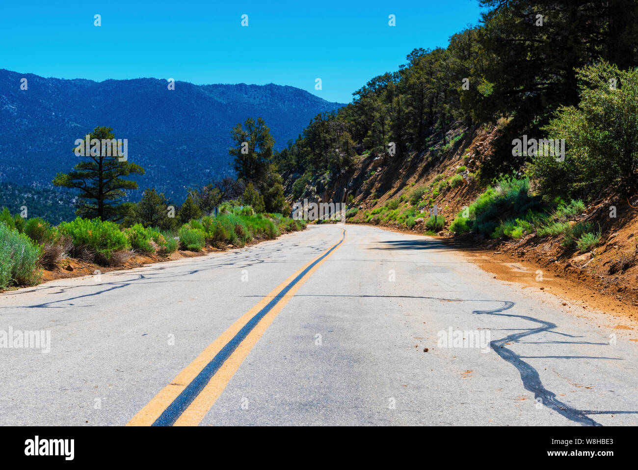 Narrow straight paved road with double yellow lines leading down hill towards mountains beyond under bright blue skies. Stock Photo