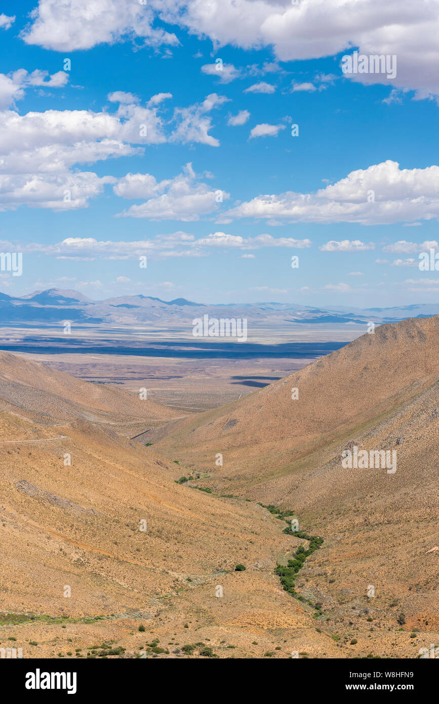 Looking down and across a desert canyon with brown mountains on each side towards valley beyond under bright blue skies with white clouds. Stock Photo