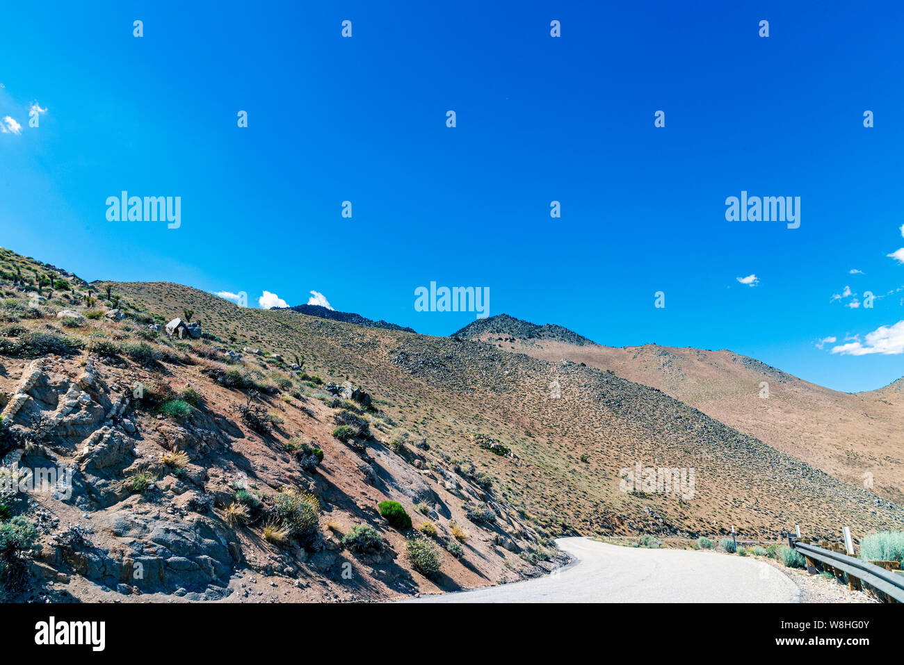 Narrow paved mountain road leading down mountain with guard rail. Desert rocky mountainside under bright blue sky. Stock Photo