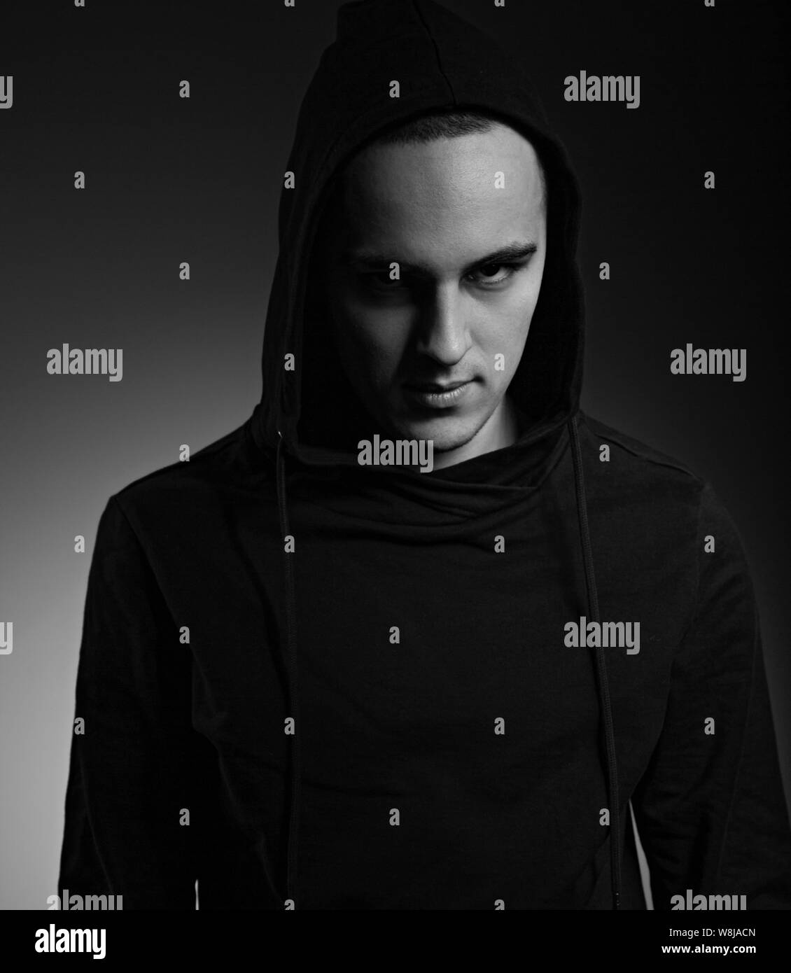 Mysterious serious man in black hoodie with hood on the head on dark background. Dangerous criminal person in dark shadow. Black and white Stock Photo