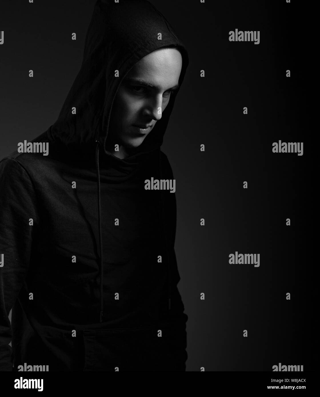 Mysterious serious man in black hoodie with hood on the head on dark background. Dangerous criminal person in dark shadow. Black and white Stock Photo