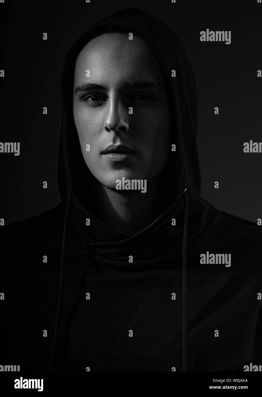 Mysterious serious man in black hoodie with hood on the head on dark background. Dangerous criminal person in dark shadow. Black and white. Art Stock Photo