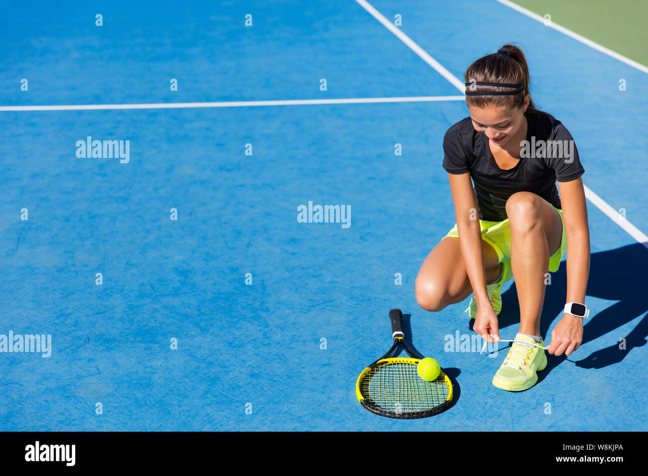 Sports woman Asian happy athlete getting ready for playing tennis tying laces of her running shoes on outdoor blue hard court. Professional player preparing for summer tournament game. Stock Photo