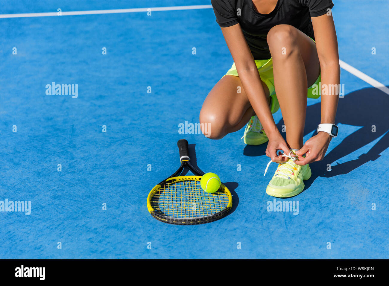 Sports woman athlete getting ready for playing a game of tennis tying laces of her running shoes lacing the shoelaces on outdoor blue hardcourt in summer. Professional player preparing for tournament. Stock Photo