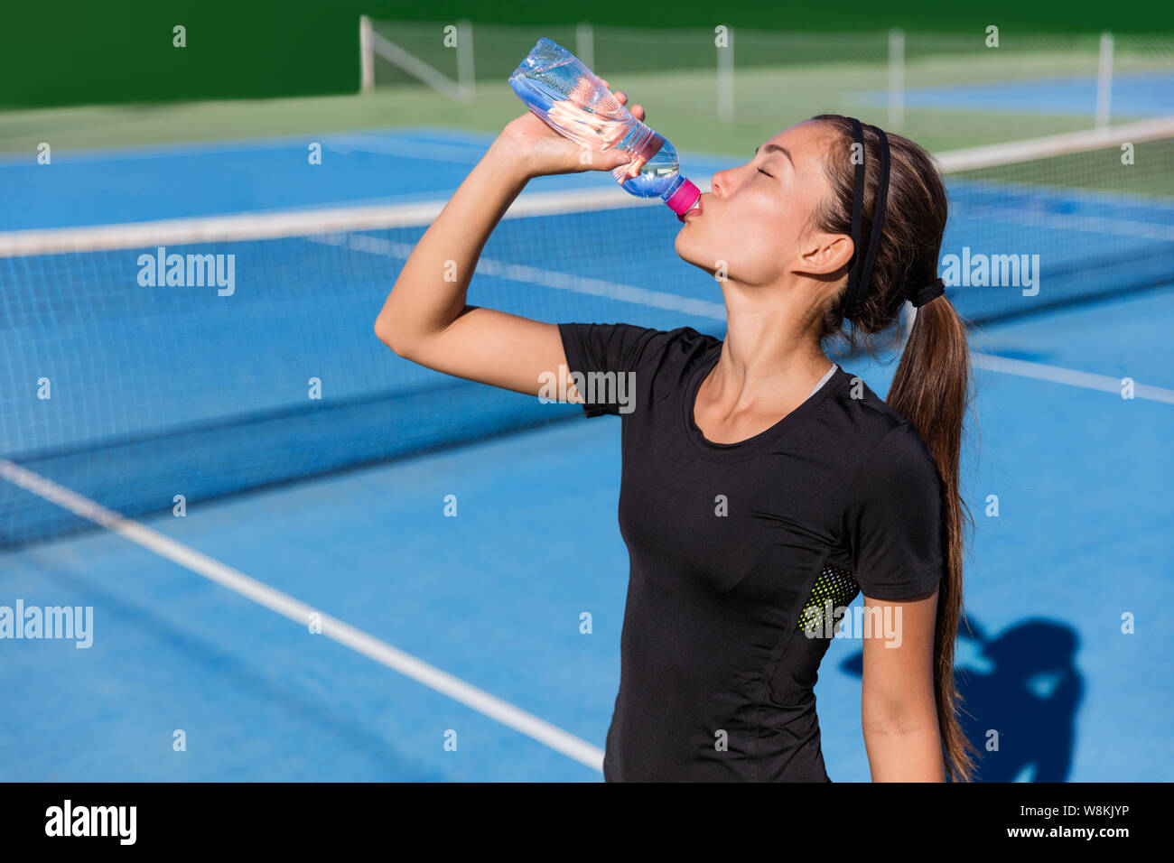 Healthy tennis player thirsty thirst quenching drinking health sports drink water plastic bottle hydrating before playing a game on blue hardcourt. Professional athlete living an active fit lifestyle. Stock Photo