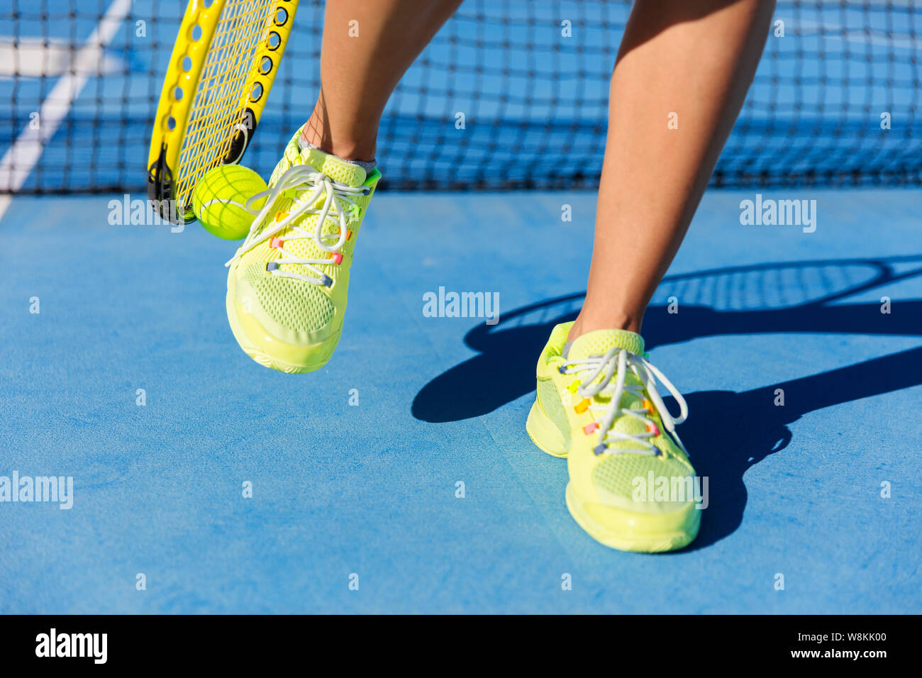 Sports athlete picking up ball with tennis racket. Female player using a technique with her running shoes to pick up during game on blue hard court. Closeup of feet, neon yellow fashion footwear. Stock Photo