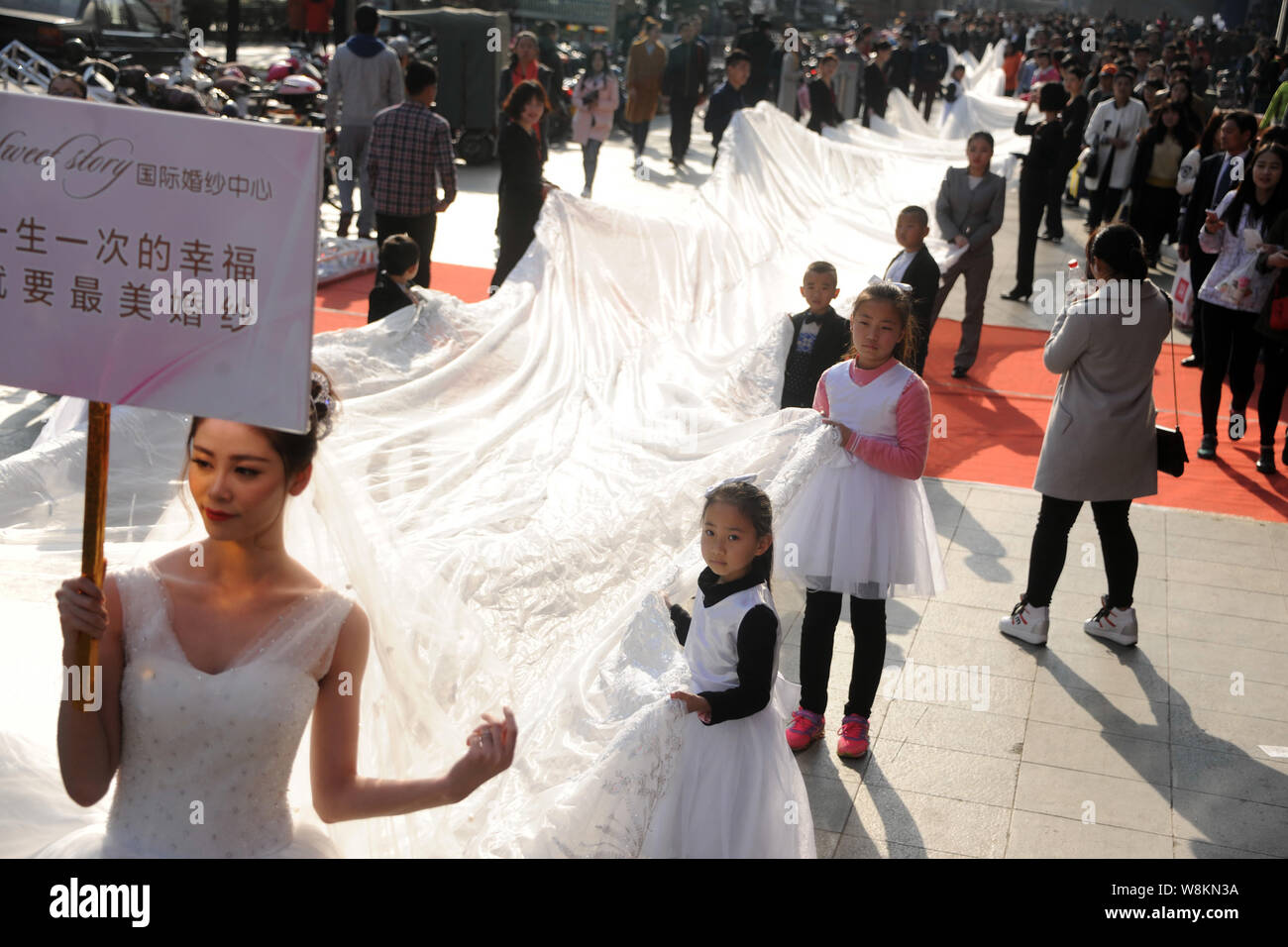 A model wearing a bridal gown with a 101-meter-long tail carried by young kids and staff members from a wedding dress company parades along a street d Stock Photo