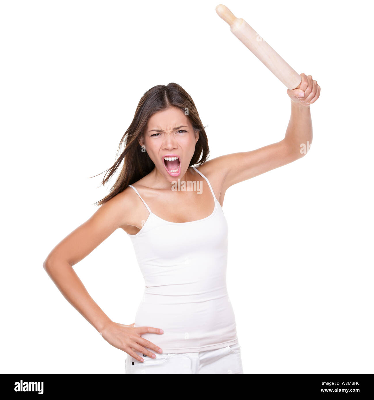 Crazy upset housewife or chef woman holding up a rolling pin in anger threatening to hit in violence, isolated on white studio background. Funny negative expression Asian girl screaming at camera. Stock Photo