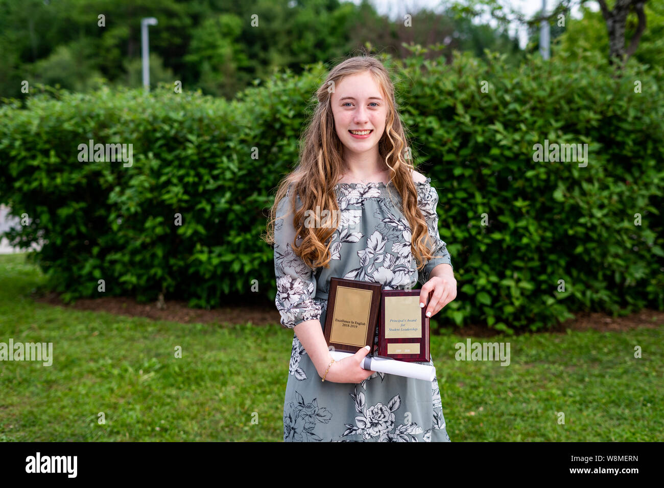 Young teen girl/Middle school student standing in front of a bush/garden after her graduation ceremony while holding a diploma and academic awards. Stock Photo