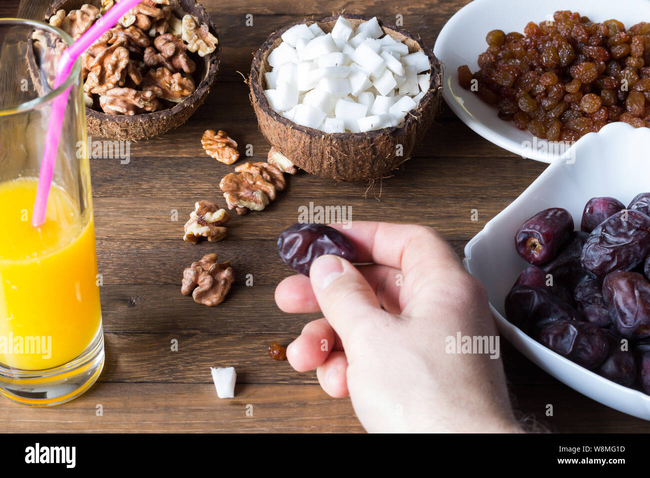 Healthy food eating process. The male hand holds a date fruit. Glass of fresh orange juice, coconut meat cubes in cup from shell, kernels of walnut, r Stock Photo