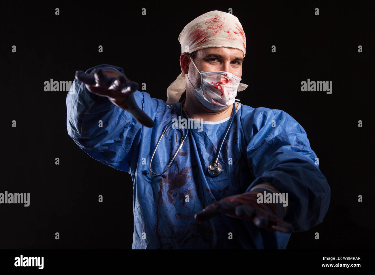 Crazy Doctor with a surgeon mask and scrubs splattered with blood for halloween. Dangerous doctor over black background. Stock Photo