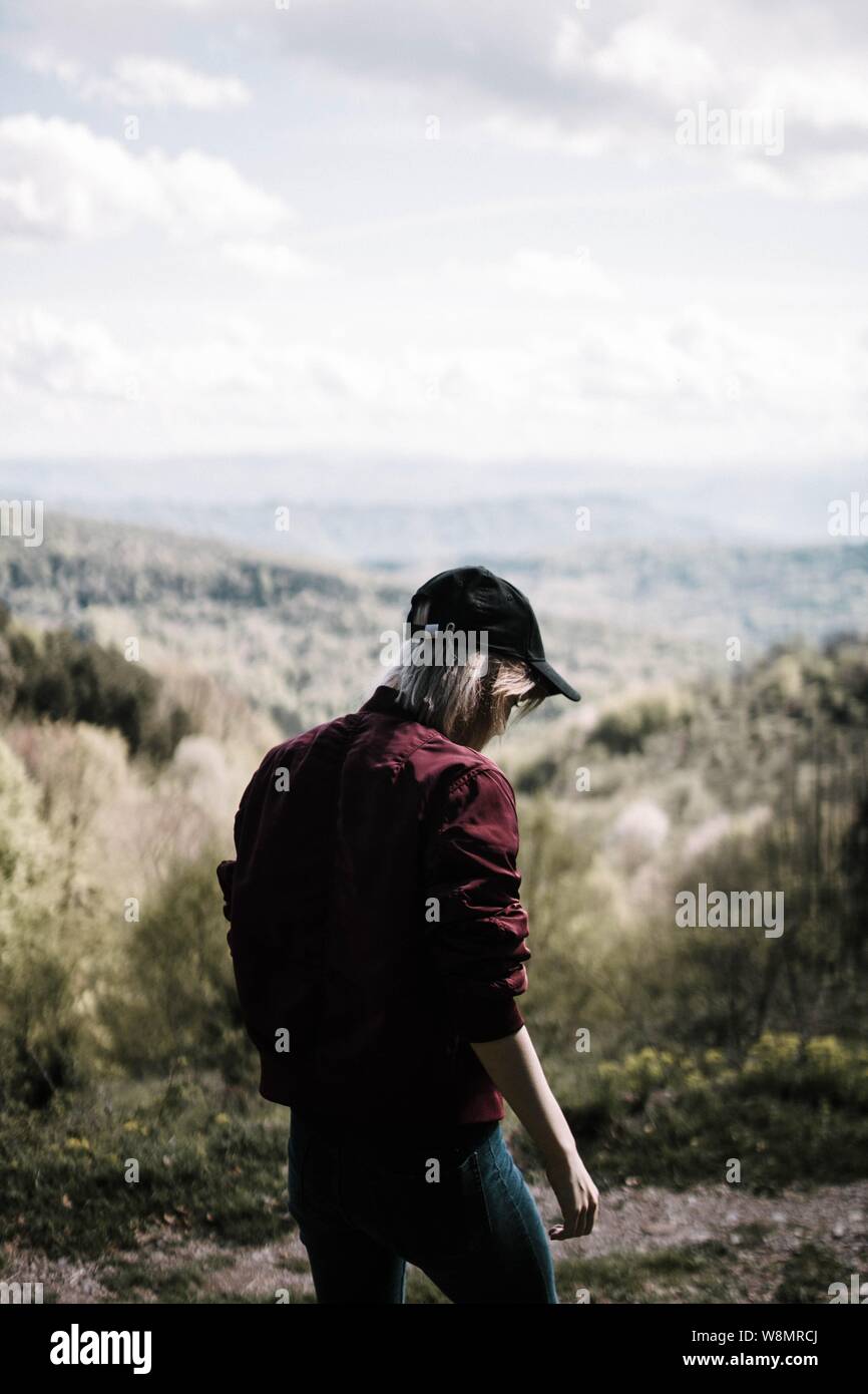 Person wearing a maroon jacket and blue jeans standing on mountain scenery Stock Photo