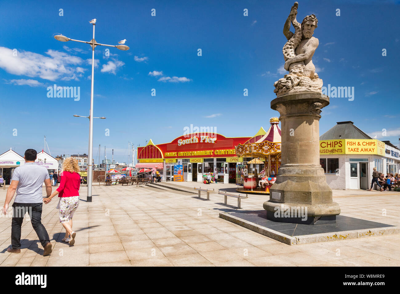 15 June 2019: Lowestoft, Suffolk, UK - A Triton statue on the promenade, with South Pier, amusements and back view of couple holding hands. Stock Photo