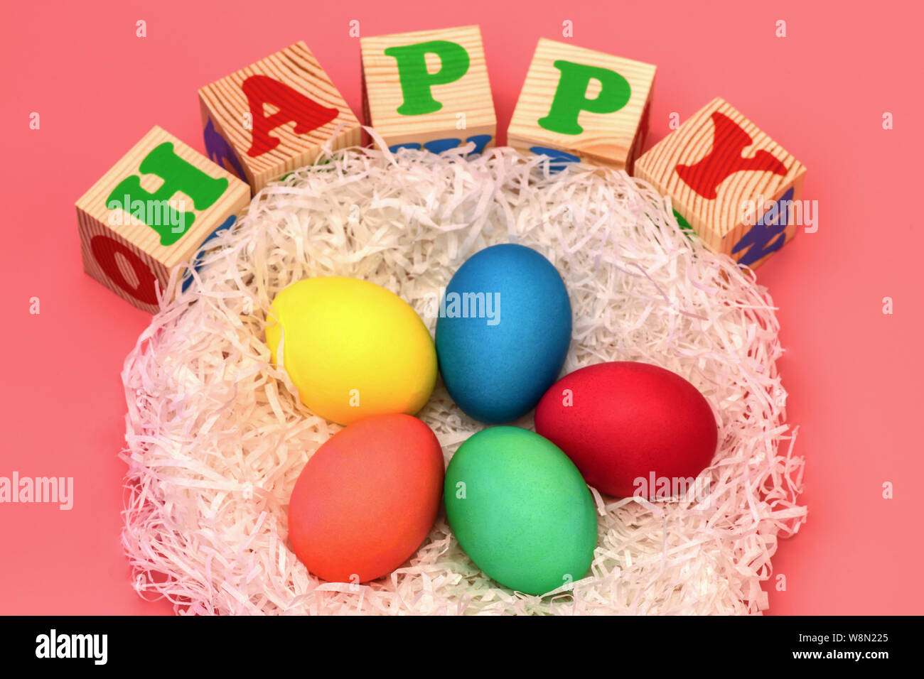 Five Easter colored eggs lie in a nest on top of cubes with the words happy on a gently pink background. Horizontal photo. Stock Photo