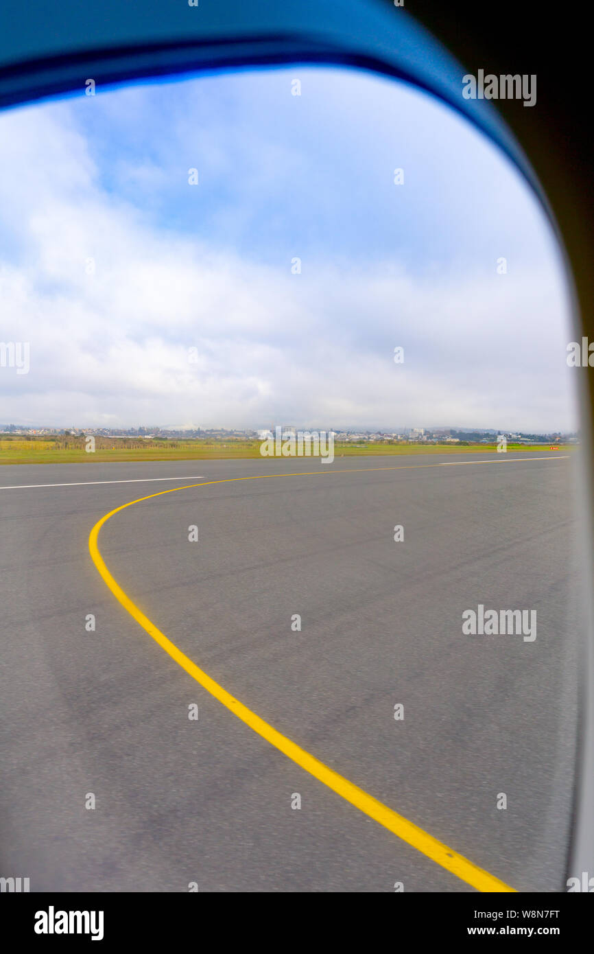 Tauranga Airport runway through plane window before take-off with long curving yellow marking and Tauranga City skyline in distance.. Stock Photo