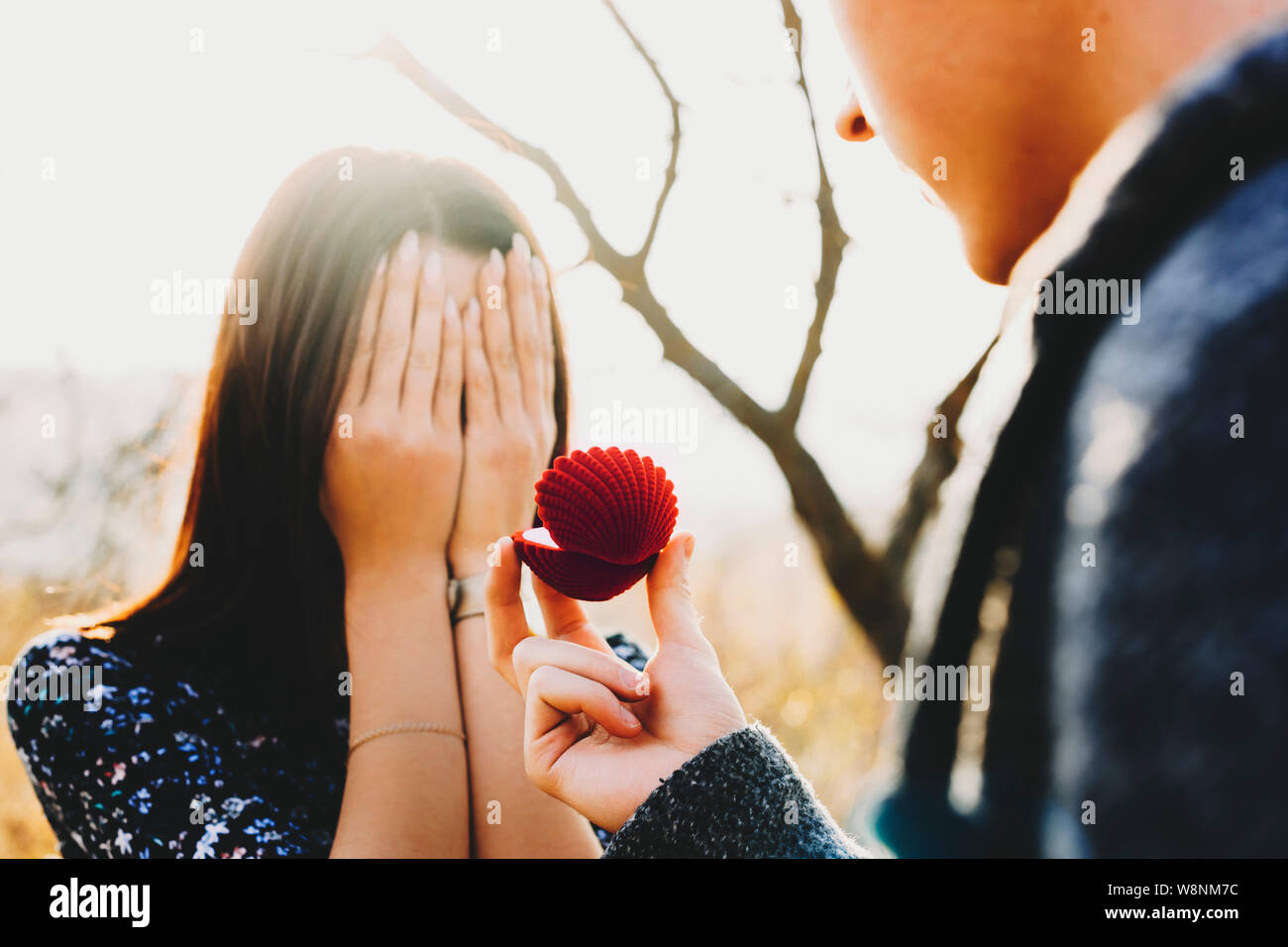 Unrecognizable woman hiding face behind hands while standing near crop male with ring box on sunny day in nature Stock Photo