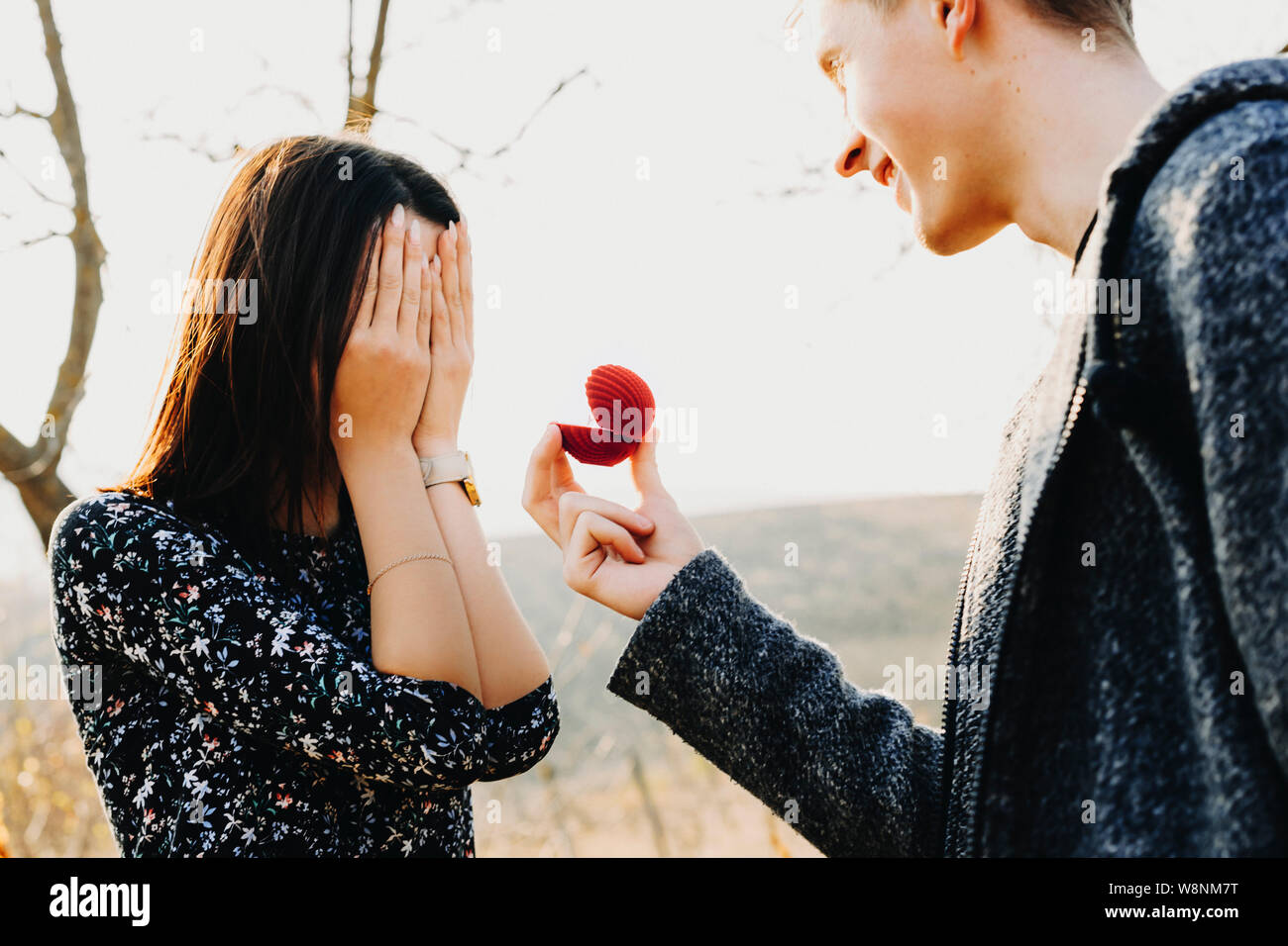 Side view of handsome young male smiling and holding small ring box while proposing to anonymous woman covering her face with hands Stock Photo