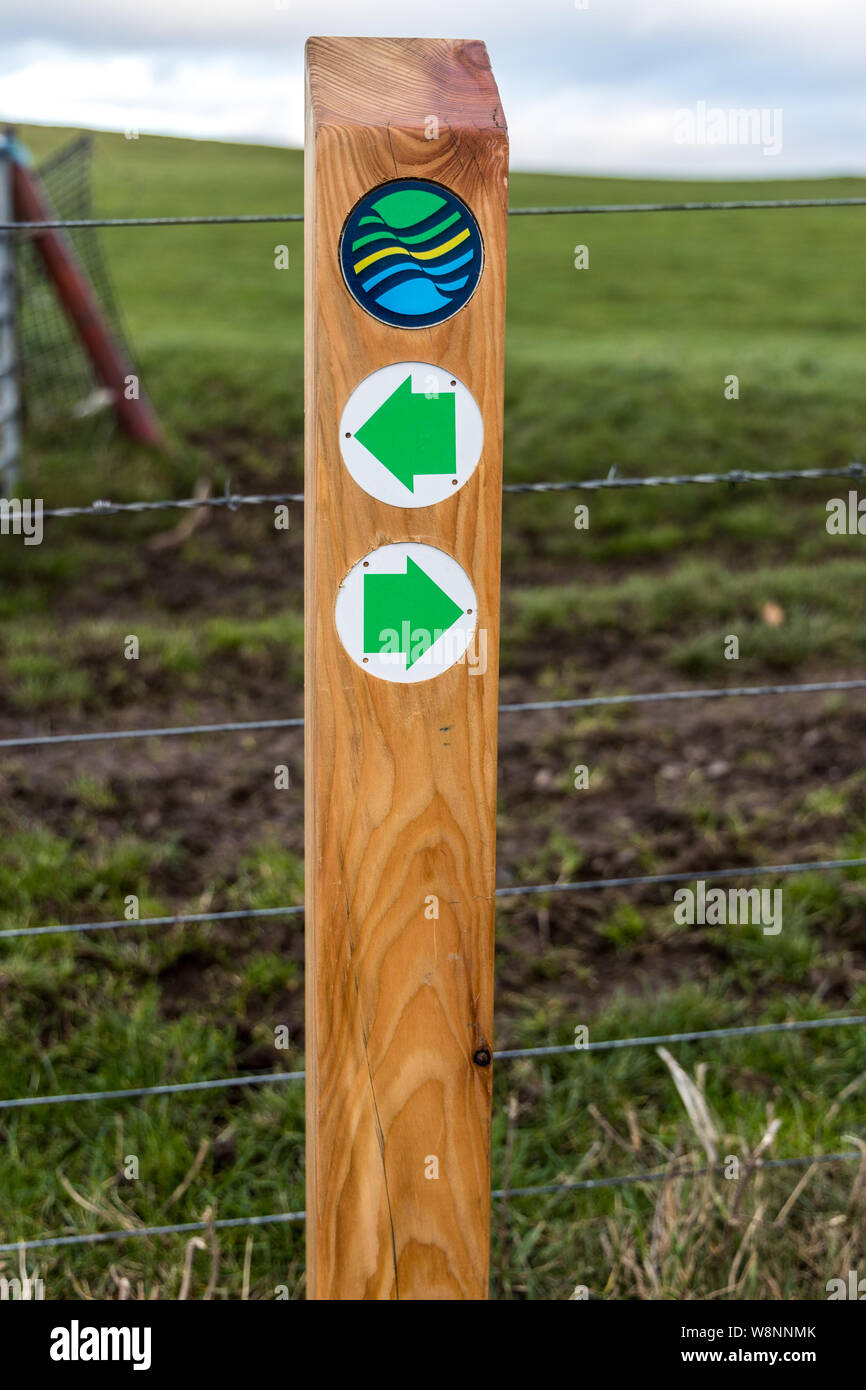 Close up of a wooden post with green arrows pointing in the direction of  the Fife Coastal Path with the logo sign above Scotland Stock Photo